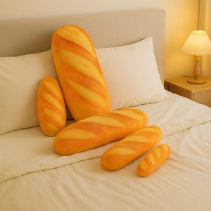 Puede incluir: Una colección de almohadas con forma de pan en varios tamaños. Las almohadas presentan un diseño realista con tonos amarillos y naranjas, imitando la apariencia de una baguette recién horneada. Están dispuestas sobre una cama con ropa de cama blanca.