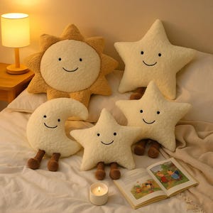 May include: A collection of plush celestial-themed pillows, including a smiling sun, moon, and three stars. The pillows are cream-colored with brown legs and smiling faces. A lit candle and an open book are also present.