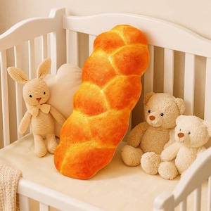 May include: A large, golden-brown challah bread-shaped cushion sits in a white cot, with a crocheted bunny and two teddy bears. The cushion has a realistic, textured look, creating a cosy nursery theme. The cot is made of white wood.