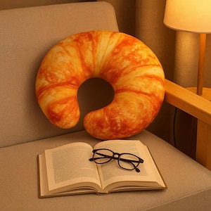 May include: A crescent-shaped pillow, coloured in shades of orange and yellow, sits on a grey chair. Below the pillow is an open book with black-framed glasses. The pillow's design mimics the texture and colour of a baked croissant.