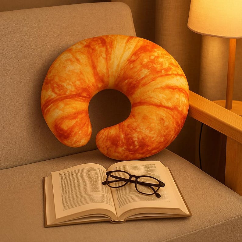 May include: A crescent-shaped pillow, coloured in shades of orange and yellow, sits on a grey chair. Below the pillow is an open book with black-framed glasses. The pillow's design mimics the texture and colour of a baked croissant.