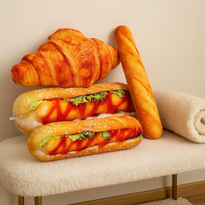 May include: A selection of plush food items, including a croissant, a baguette, and two hot dogs with sesame seed buns, lettuce, and ketchup. The items are arranged on a white bench, creating a playful and whimsical display.