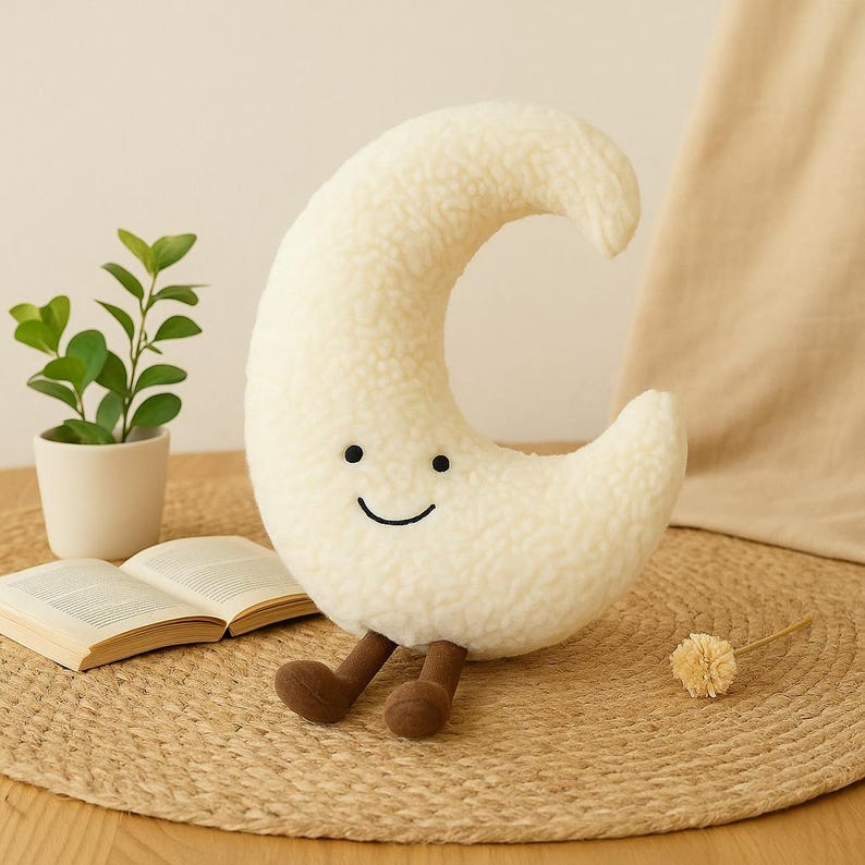 May include: A plush, cream-colored crescent moon pillow with a smiling face and brown legs. The pillow is on a woven mat, next to a small potted plant and an open book. The moon has a soft, textured surface.