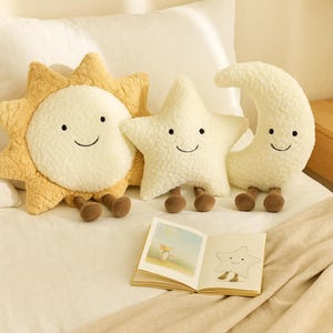 May include: Three plush celestial-themed pillows: a smiling sun, star, and crescent moon. The sun is yellow and cream, the star and moon are cream. Each has brown feet. An open book with a star illustration rests in front.
