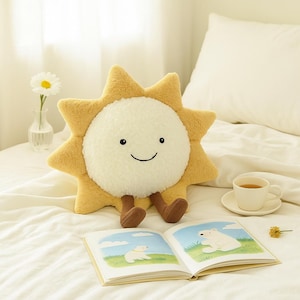 May include: A plush sun-shaped pillow with a smiling face, yellow rays, and brown legs. The pillow is on a bed with a book open to a colorful illustration. A cup of tea and a small vase with a flower are also on the bed.