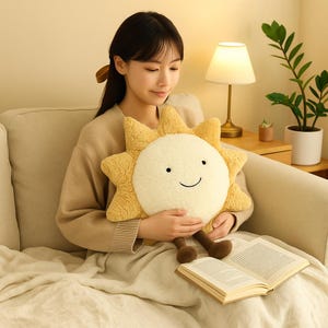 May include: A plush, smiling sun-shaped pillow with a cream-colored face and yellow rays. The pillow has brown legs and is being held. An open book rests on a beige blanket. The background includes a lamp and a potted plant.