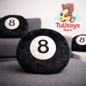 Op de afbeelding: Zwart pluche rond kussen met een witte cirkel en het nummer 8 in het midden. Het kussen ligt op een grijze ondergrond. De afbeelding toont ook twee andere soortgelijke kussens en een cartoon berenlogo.