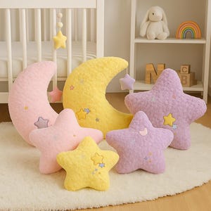 Puede incluir: Una colección de decoración de guardería de felpa con una luna creciente rosa, una luna creciente amarilla y almohadas en forma de estrella en rosa, amarillo y lavanda. Las almohadas tienen detalles bordados de estrellas y lunas.