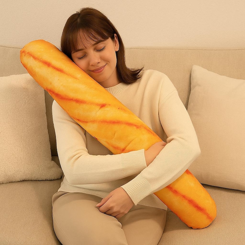 May include: A large, plush baguette-shaped pillow. The pillow is a golden-yellow colour with reddish-orange stripes, resembling a freshly baked loaf. It's designed to be soft and huggable, suitable for home decoration and comfort.