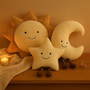 May include: A plush set of celestial-themed pillows, including a smiling sun, star, and crescent moon. The pillows are cream-colored with brown accents and legs, arranged on a wooden surface. A lit candle and a decorative cloth are also present.
