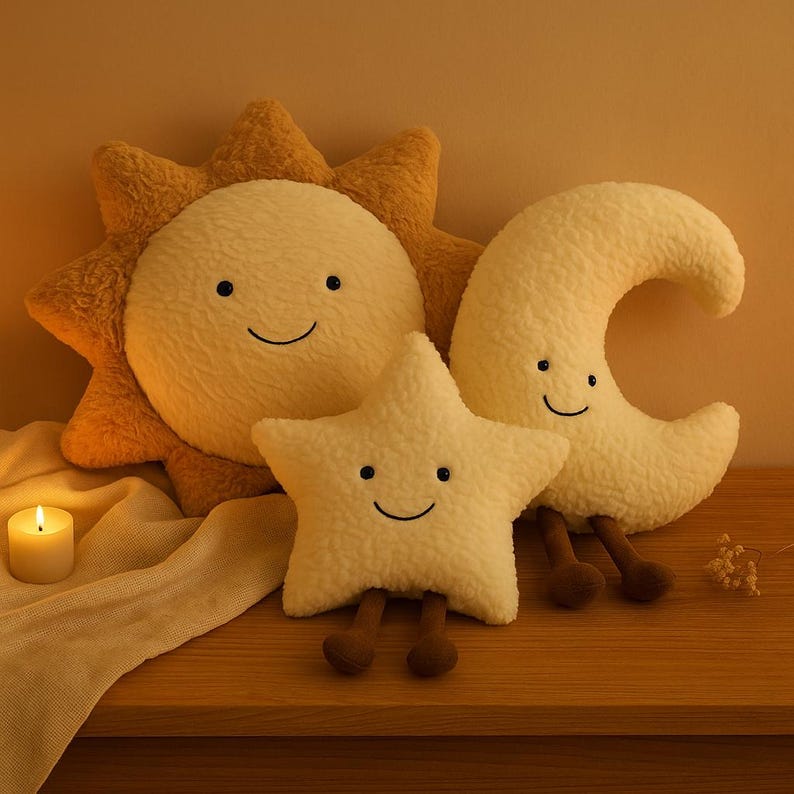 May include: A plush set of celestial-themed pillows, including a smiling sun, star, and crescent moon. The pillows are cream-colored with brown accents and legs, arranged on a wooden surface. A lit candle and a decorative cloth are also present.