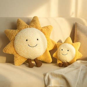 May include: Two plush sun-shaped pillows with smiling faces. The larger pillow has brown legs. The suns are yellow with a white center. The pillows are on a beige couch with a white blanket.