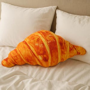 May include: A large, croissant-shaped cushion in shades of golden brown and orange, placed on a white bed. The cushion's design mimics the texture and appearance of a freshly baked croissant, with detailed ridges and a realistic colour gradient.