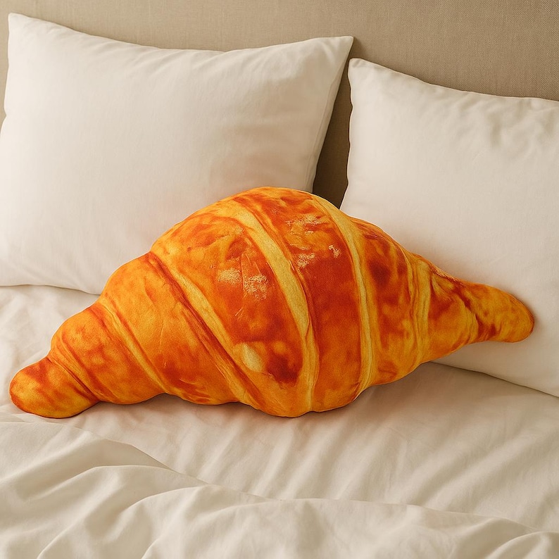 May include: A large, croissant-shaped cushion in shades of golden brown and orange, placed on a white bed. The cushion's design mimics the texture and appearance of a freshly baked croissant, with detailed ridges and a realistic colour gradient.