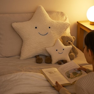 May include: Two white star-shaped plush pillows with smiling faces and a small brown teddy bear are on a bed. The larger star pillow has a textured surface. The scene is lit by a warm bedside lamp.