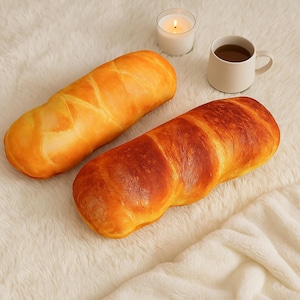 May include: Two bread-shaped cushions in shades of golden brown and tan sit on a white, fluffy surface. A lit candle and a mug of tea are also present. The cushions mimic the appearance of freshly baked bread, ideal for home decor.