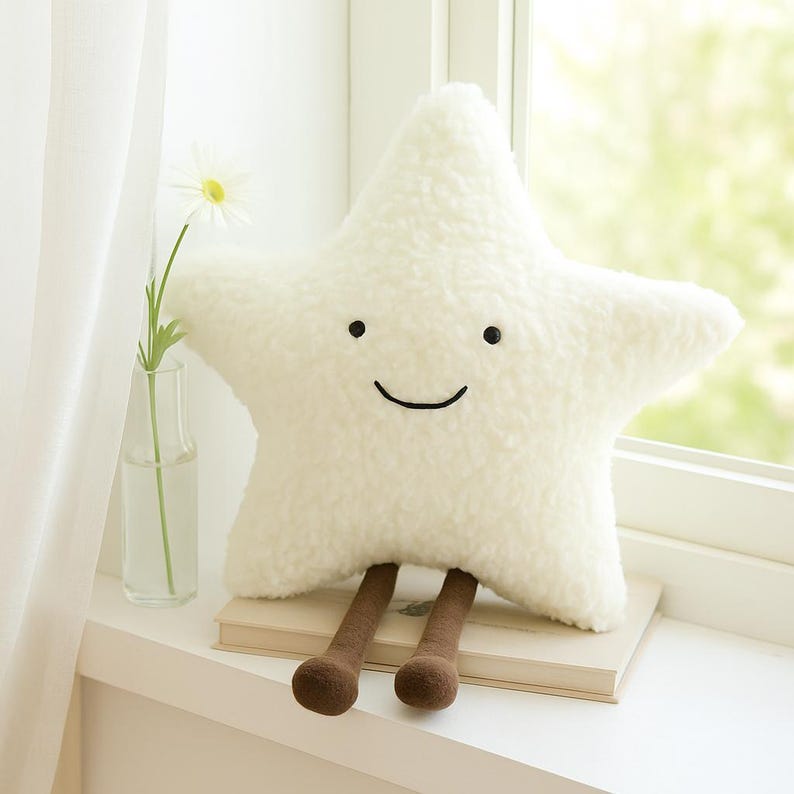 May include: A plush, white star-shaped pillow with a smiling face and brown legs. The pillow is made of a soft, textured material and sits on a stack of books. A daisy in a glass vase is in the background.