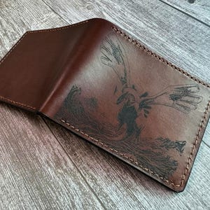 May include: A dark brown leather bifold wallet featuring a detailed black laser-engraved illustration of a winged creature with outstretched wings. The wallet is constructed with visible thick stitching along the edges and a smooth, polished finish.