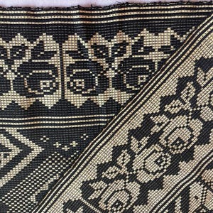 Può includere: Tessuto nero e beige tessuto con un motivo floreale e geometrico ripetuto. Il tessuto è piegato, rivelando il design e la trama complessi. Il motivo include fiori stilizzati e forme a diamante.