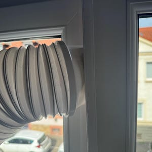 Puede incluir: Un tubo de escape blanco, flexible y de estilo acordeón para un aire acondicionado portátil, ajustado de forma segura en un marco de ventana blanco. El tubo presenta una serie de crestas circulares y se extiende horizontalmente desde la ventana.