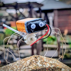 May include: A DIY robotic spider constructed from a 9-volt battery, wires, and a small motor. The battery is orange and black, with the text "10000" visible. The spider's legs are made of silver wire, and it is perched on a textured rock.