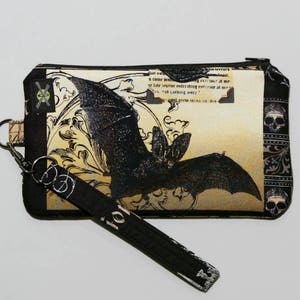 May include: A black and white bat with intricate details on a beige background. The bat is surrounded by floral designs and the text "that we never..." is visible. The pouch has a black strap with a silver clasp and a skull design on the side.