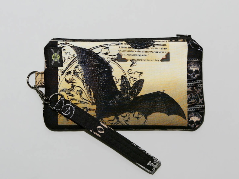 May include: A black and white bat with intricate details on a beige background. The bat is surrounded by floral designs and the text "that we never..." is visible. The pouch has a black strap with a silver clasp and a skull design on the side.