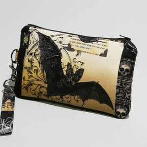 May include: A black and beige zippered pouch with a black bat graphic and a black and white floral pattern. The pouch has a black strap with a silver clasp.