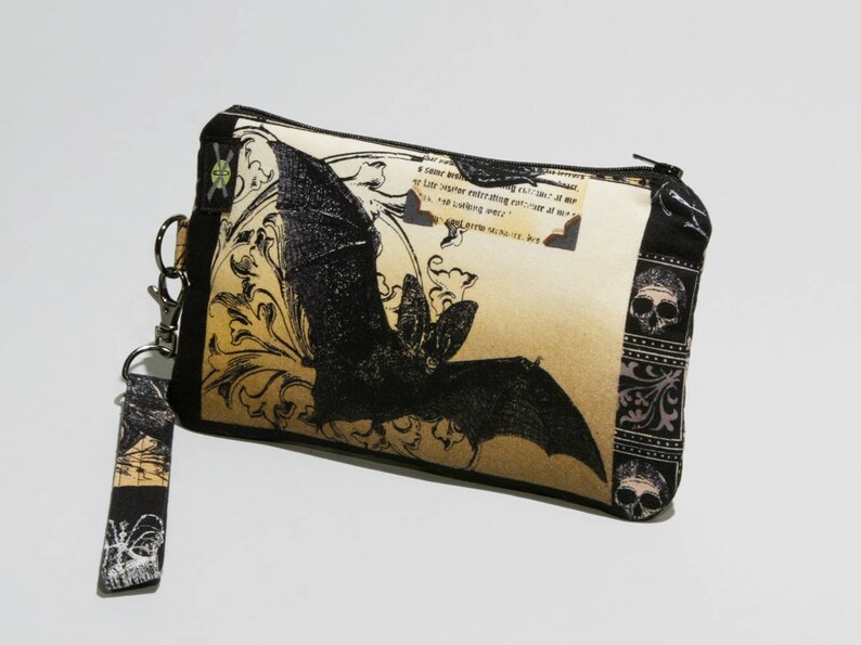 May include: A black and beige zippered pouch with a black bat graphic and a black and white floral pattern. The pouch has a black strap with a silver clasp.