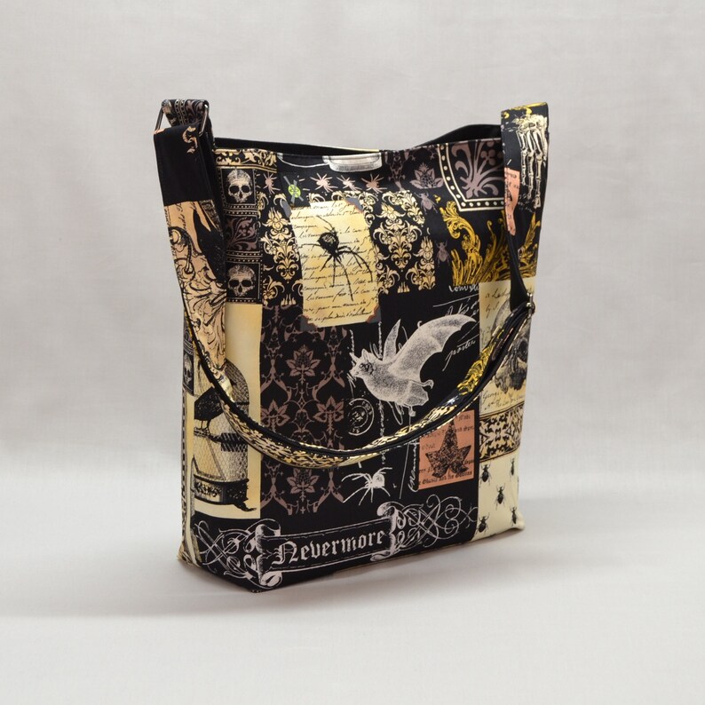 Pode incluir: Uma sacola preta com tema de Halloween. A sacola apresenta um design de patchwork com imagens de morcegos, aranhas, caveiras e a palavra "Nevermore". A sacola tem uma al&ccedil;a de ombro e formato retangular.