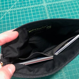 May include: Black zippered pouch with a black fabric interior. The pouch has a small pocket on the inside and is open to reveal several credit cards inside. The pouch has a yellow label with the text "Working Class Punk" on it.