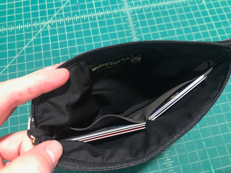 May include: Black zippered pouch with a black fabric interior. The pouch has a small pocket on the inside and is open to reveal several credit cards inside. The pouch has a yellow label with the text "Working Class Punk" on it.