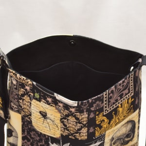 Pode incluir: Bolsa tote preta com um padr&atilde;o tem&aacute;tico de Halloween. A bolsa apresenta um design de patchwork com caveiras, aranhas e volutas ornamentadas. O interior &eacute; preto e a bolsa tem um fecho magn&eacute;tico de press&atilde;o. A bolsa tem uma al&ccedil;a de ombro.