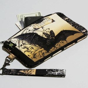 May include: A black and gold fabric wristlet with a bat design and a zipper closure. The wristlet has a detachable strap and is made of a soft, durable fabric.
