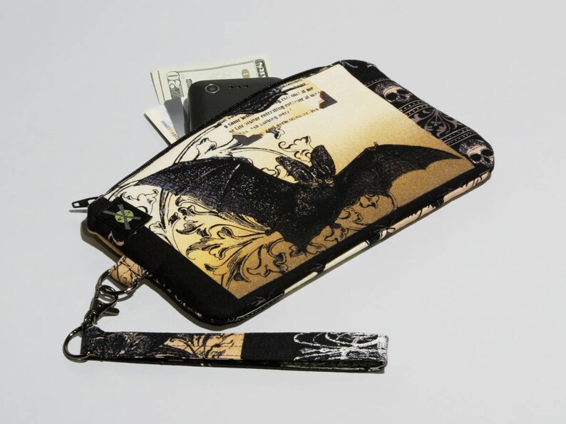 May include: A black and gold fabric wristlet with a bat design and a zipper closure. The wristlet has a detachable strap and is made of a soft, durable fabric.