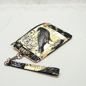 Puede incluir: Una bolsa con cremallera negra y dorada con un cuervo negro y un dise&ntilde;o floral. La bolsa tiene una correa negra y dorada.