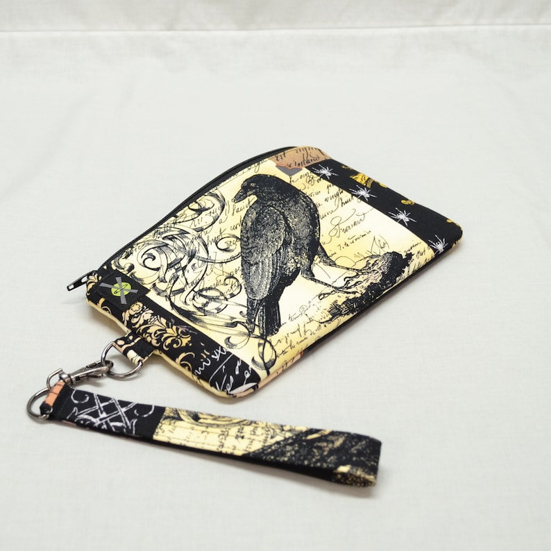 Puede incluir: Una bolsa con cremallera negra y dorada con un cuervo negro y un dise&ntilde;o floral. La bolsa tiene una correa negra y dorada.