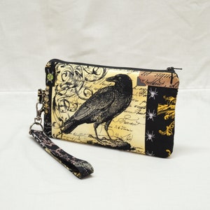Puede incluir: Una bolsa con cremallera negra y amarilla con un cuervo negro y un dise&ntilde;o floral. La bolsa tiene una correa negra con un cierre plateado.