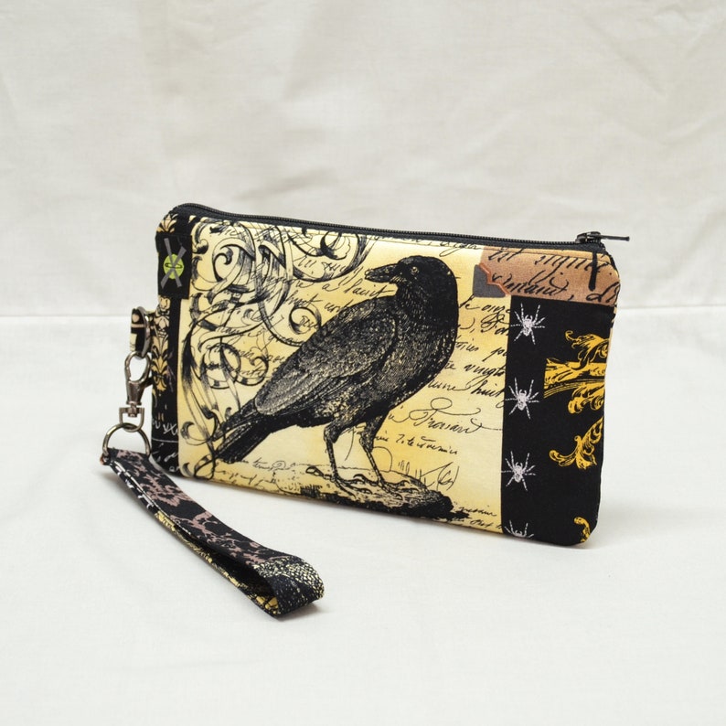 Puede incluir: Una bolsa con cremallera negra y amarilla con un cuervo negro y un dise&ntilde;o floral. La bolsa tiene una correa negra con un cierre plateado.