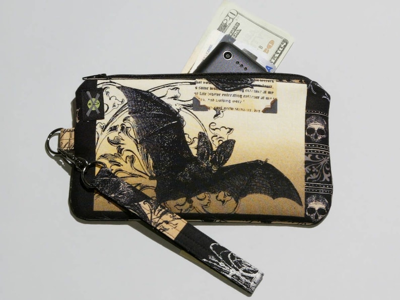 May include: A black and beige zippered pouch with a bat graphic and a skull pattern. The pouch has a wrist strap and a zippered pocket on the back.