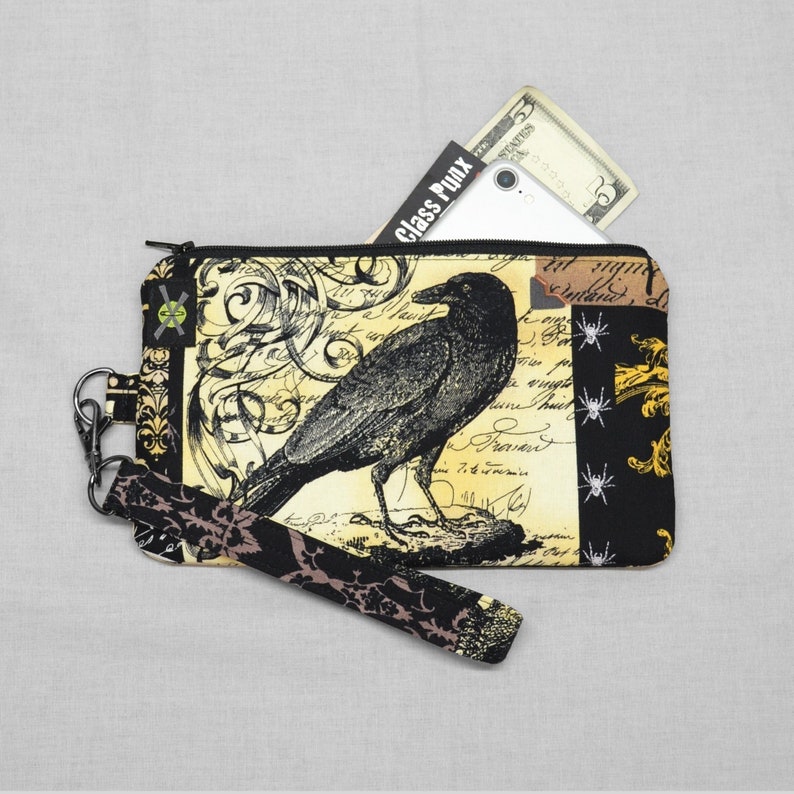 Puede incluir: Bolsa negra con cremallera con un cuervo negro y un dise&ntilde;o floral dorado. La bolsa tiene una correa negra y dorada y el texto "Class Punk" es visible en una tarjeta blanca dentro de la bolsa.