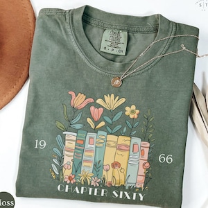 May include: A moss green t-shirt featuring a floral and book design. The graphic includes colorful flowers, a stack of books, and the text "CHAPTER SIXTY" with the numbers "19" and "66". A gold necklace is draped across the shirt.