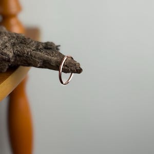 May include: A delicate, thin rose gold ring. The ring is displayed on a piece of textured, dark brown wood, set against a soft gray background. The ring's simple design highlights its elegant, minimalist style.