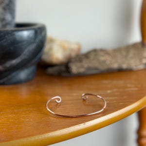 May include: A delicate copper-colored cuff bracelet with curled ends rests on a wooden surface. The bracelet's simple design and warm hue create a minimalist aesthetic. The bracelet is a stylish accessory.