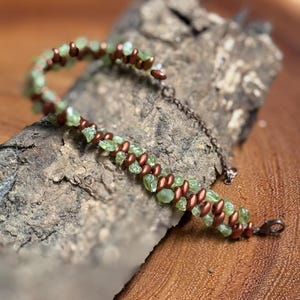 May include: A handmade bracelet featuring light green gemstone beads and copper-colored metal beads. The bracelet is displayed on a piece of wood, showcasing its natural and earthy aesthetic. The clasp is a small, copper-colored hook.