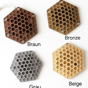Op de afbeelding: Vier zeshoekige onderzetters in bruin, brons, grijs en beige. Elke onderzetter heeft een honingraatpatroon en een klein gaatje om op te hangen. De kleuren zijn gelabeld met "Braun", "Bronze", "Grau" en "Beige".
