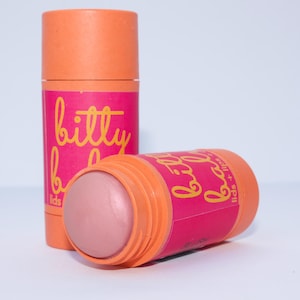 Peut inclure: Deux tubes de baume pour les lèvres et les paupières. Les tubes sont orange et rose, avec l'inscription "bitty balm" en jaune. Un tube est debout, l'autre est couché sur le côté, révélant le baume rose. Le fond est blanc.