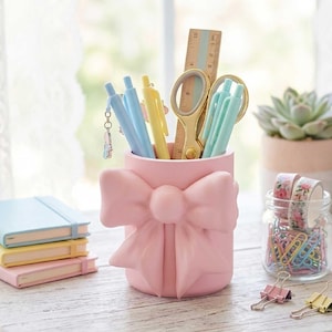 Op de afbeelding: Een roze strikvormige bureau-organizer bevat pennen, een schaar en een liniaal. Een klein glazen potje bevat kleurrijke paperclips en bloemen washi tape. De afbeelding bevat ook een vetplant, notitieboekjes en de tekst "STL 3MF 3D PRINT FILE".
