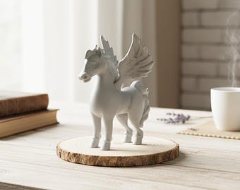 May include: A white ceramic Pegasus figurine with detailed wings, standing on a round wooden slice. The mythical creature is posed with its head up, showcasing its elegant form. The background includes a window and a cup.