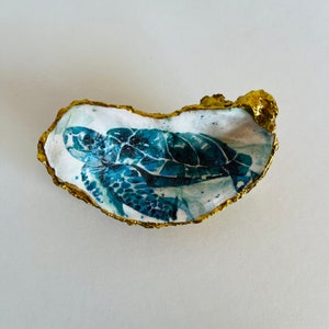 May include: A decorative oyster shell with a gold-painted edge. The interior features a watercolor painting of a sea turtle in shades of blue and green. The shell is set against a white background, highlighting the artwork.